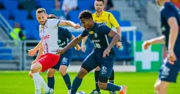 Piłkarze walczą o piłkę na boisku. Jeden w białej koszulce, drugi w granatowej. Sędzia obserwuje grę. To fragment meczu z ligi francuskiej.