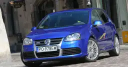 Błękitny Golf R32 z felgami ze stopów lekkich, zaparkowany na brukowanej uliczce przed kawiarnią.