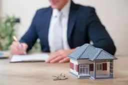 Model domu i klucze na biurku, a w tle mężczyzna w garniturze podpisuje dokumenty. Dowiedz się, jak ubezpieczyć mieszkanie.