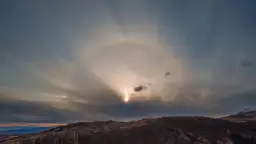 Słońce otoczone halo na tle chmur nad wzgórzem.