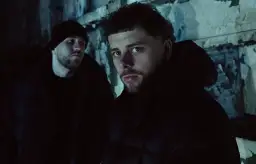 Dwóch mężczyzn w ciemnych kurtkach i czapce na tle zniszczonej ściany.