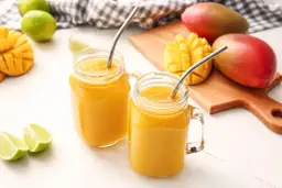 Dwa orzeźwiające drinki z mango w słoikach z metalowymi słomkami, obok świeżych mango i limonek.