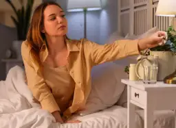 Młoda kobieta w łóżku sięga po lampkę, zastanawiając się, czy warto obniżać temperaturę na noc. Obok budzik i szklanka wody.