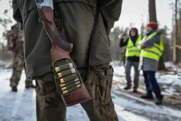 Myśliwy w lesie, z bronią i amunicją w pasie. W tle inni uczestnicy polowania w kamizelkach odblaskowych.