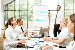 Zespół omawia strategię marketing internetowy. Prezentacja danych na tablicy, ludzie przy stole z dokumentami i laptopem.