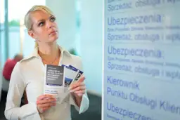 Kobieta z ulotkami zastanawia się nad przedłużeniem OC przed terminem. Na tablicy widnieje napis "Ubezpieczenia".