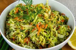 Surówka z kapusty i marchewki z koperkiem w białej misce, obok plasterek cytryny.