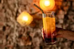 Dłoń trzymająca szklankę z pomarańczowym napojem i lodem, na tle ceglanej ściany i świateł.