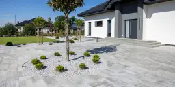 Nowoczesny dom z szarym podjazdem z kostki, otoczony zadbanym ogrodem z drzewami i krzewami.