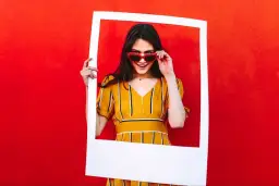 Uśmiechnięta kobieta w okularach w kształcie serca trzyma białą ramkę na tle czerwonej ściany.