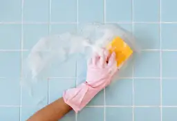 Ręka w różowej rękawiczce myje gąbką niebieskie kafelki łazienkowe z pianą.