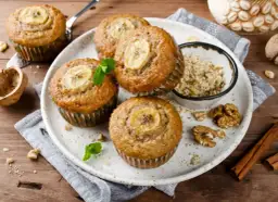 Pięć pysznych muffinek bananowych z plasterkiem banana na wierzchu, udekorowane miętą. Obok miseczka z orzechami i laska cynamonu.