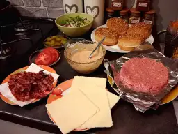 Składniki do przygotowania idealnego cheeseburgera: bułki, surowe mięso, ser, bekon, pomidory, ogórki i sos.
