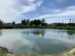 Spokojny staw z odbijającymi się chmurami, otoczony zielenią i zabudowaniami.