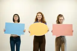 Trzy dziewczyny z kolorowymi dymkami. Jedna z nich zastanawia się, jak nauczyć się mówić po angielsku.