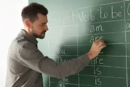 Nauczyciel pisze na tablicy odmianę czasownika "to be". Nauka angielskiego na poziomie C1 to cel wielu uczniów.