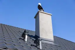 Komin na dachu pokrytym ciemną dachówką, z widocznym obrobieniem blachą. Wiatrownica na kominie.