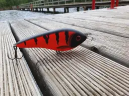 Czerwono-czarna przynęta wędkarska na szczupaka leżąca na drewnianym pomoście.