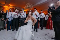 Panna młoda siedzi na krześle, śmiejąc się, otoczona przez weselnych gości i kamerzystę.