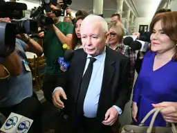 Jarosław Kaczyński w garniturze, otoczony przez dziennikarzy z mikrofonami. Wzrost i waga polityka są widoczne w jego postawie.