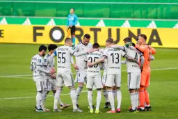 Piłkarze Legii Warszawa w białych strojach stoją w kółku na boisku, przytulając się.