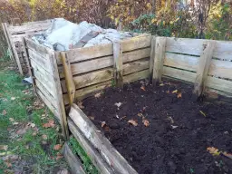 Drewniany kompostownik w ziemi, częściowo przykryty plandeką. Widać jesienne liście i trawę.