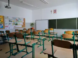 Pusta sala lekcyjna z rzędami krzeseł i biurek, tablicą i dziecięcymi rysunkami na ścianie.