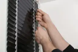 Dłonie regulują czarną roletę, pociągając za białą taśmę.