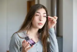 Dziewczyna wykonuje makijaż oczu, aplikując cień do powiek. Dba o wygląd twarzy, używając paletki z fioletowymi odcieniami.