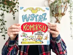 Okładka książki "Mistrz Komiksu" z rysunkami postaci i słońca, trzymana przez ręce.