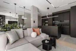 Nowoczesny salon z kuchnią, gdzie oświetlenie wpuszczane tworzy elegancką atmosferę. Szara sofa, czarne meble kuchenne i marmurowe detale.