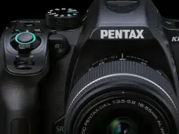 Zbliżenie na czarną lustrzankę Pentax KM z obiektywem 18-55mm. Widoczne pokrętła, przyciski i logo producenta.