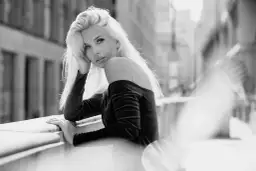 Elegancki portret w plenerze. Kobieta z blond włosami opiera się o balustradę, patrząc w obiektyw.