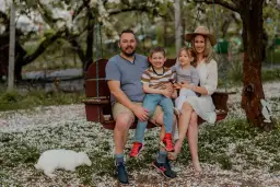 Rodzina na huśtawce pod kwitnącym drzewem. Tata z synem na kolanach, mama z córką. W tle biały królik. Idealne cytaty o tacie.