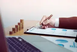 Ręka pisze na dokumentach z wykresami, obok stosy monet. Sąd może żądać wyciągu z konta w sprawach finansowych.