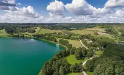 Widok z lotu ptaka na turkusowe jezioro, zielone wzgórza i lasy, z kilkoma domami.