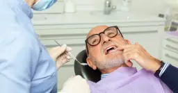 Dentysta w masce i rękawiczkach bada uśmiech starszego mężczyzny w okularach.