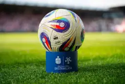 Piłka do piłki nożnej na niebieskim postumencie z logo Ekstraklasy na tle stadionu.