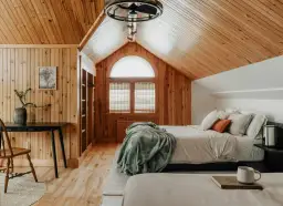 Dormitorio con paredes y techo de madera, cama con mantas y almohadas, ventana arqueada y escritorio.