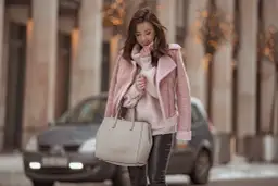 Kobieta w różowym kożuszku, swetrze i skórzanych spodniach trzyma torebkę.