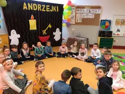 Dzieci na andrzejkach w przedszkolu, siedzą w kółku. W tle dekoracje z napisem "ANDRZEJKI", świece i duchy.