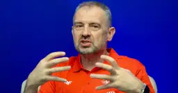 Mężczyzna w pomarańczowej koszulce z logo, gestykuluje rękami na tle niebieskiego tła.