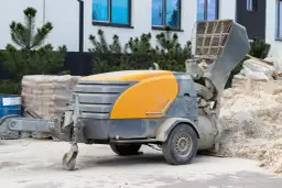 Mobilna maszyna do betonu, gotowa do pracy nad posadzkami z miksokreta. Obok piasek i materiały budowlane.