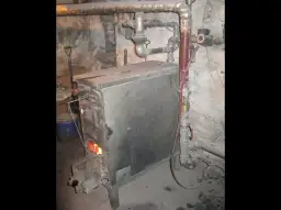 Stary piec węglowy z płomieniami w palenisku, otoczony rurami i narzędziami. Trudno ocenić, ile waży piec węglowy, ale jest solidny.