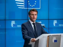 Emmanuel Macron w garniturze przemawia za mównicą na tle niebieskiej ściany z logo Unii Europejskiej.