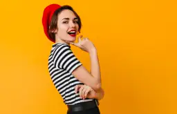 Kobieta w czerwonym beretce i pasiastym sweterku, z ustami pomalowanymi na czerwono, uśmiecha się na tle pomarańczowym. To kwintesencja paryskich kolorów.