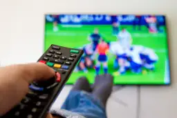Dłoń trzymająca pilot do telewizora, na ekranie mecz piłki nożnej.