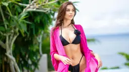 Kobieta w czarnym bikini i różowym narzutce pozuje na tle tropikalnej roślinności i oceanu. Wygląda jak z **ola hotel paradise**.