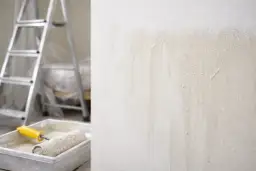 Ślady po malowaniu pod światło, widoczne na chropowatej ścianie. Obok wałek w kuwecie i drabina.