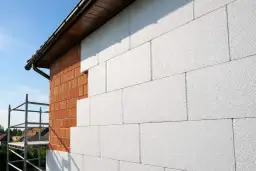 Ocieplenie ścian zewnętrznych domu w trakcie prac. Widoczna ceglana ściana i białe płyty izolacyjne. W tle rusztowanie.
